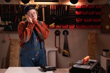 Kadın katılımcı atölye sahnesinde atölye kıyafetleri giyer duvarda asılı duran aletlerle uyku jesti yapar. Bayan marangoz gece geç saatlerde otantik garajda. Boşluğu kopyala