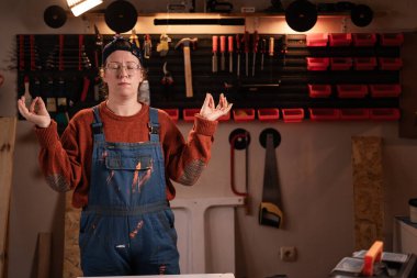 Kadın katılımcı atölye sahnesinde atölye kıyafetleri giyer duvarda asılı duran aletlerle yoga om jestinde el ele tutuşur rahatla meditasyon yapar. Bayan marangoz gece geç saatlerde otantik garajda. Boşluğu kopyala