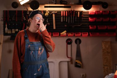 Kadın katılımcı atölye sahnesinde atölye kıyafetleri giyer. Duvarda asılı duran aletler kameraya poz verirken esniyordur. Bayan marangoz gece geç saatlerde otantik garajda. Boşluğu kopyala