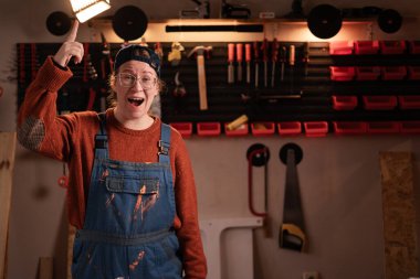 Kadın katılımcı atölye sahnesinde atölye kıyafetleri giyer. Duvarda asılı duran aletler iyi bir fikir olduğunu gösterir. Bayan marangoz gece geç saatlerde otantik garajda. Boşluğu kopyala