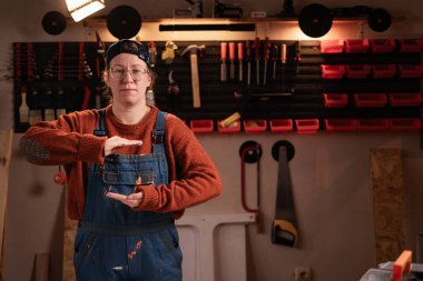 Kadın katılımcı atölye sahnesinde atölye kıyafetleri giyer, duvarda asılı aletlerle büyük bir kutu ve el hareketleriyle büyük bir ürün gösterir. Bayan marangoz gece geç saatlerde otantik garajda. Boşluğu kopyala