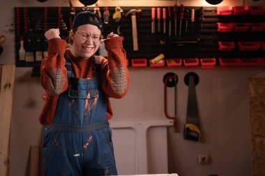 Kadın katılımcı atölye sahnesinde atölye kıyafetleri giyer duvarda asılı aletlerle kazanan jestler yaparak yumruklarını sıkmanı kutlar. Bayan marangoz gece geç saatlerde otantik garajda. Boşluğu kopyala