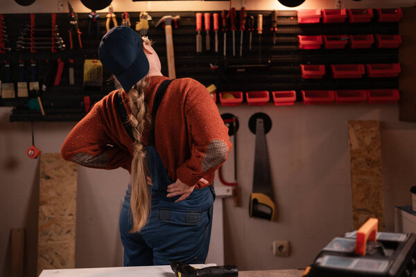 Health Problem Concept. Woman joiner wears workwear in workshop scene with tools hanging on wall suffering from acute backpain. female carpenter late evening in authentic garage. copy space