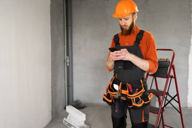 İnşaat işçisi sakallı adam inşaat alanında akıllı telefon kullanıyor ya da ev tamiri yapıyor. Profesyonel inşaatçı içeride. Boşluğu kopyala
