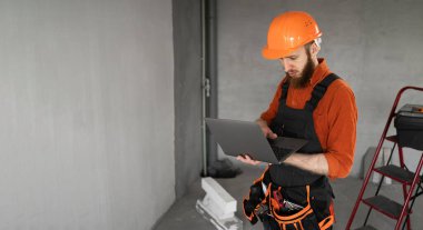 İnşaat işçisi sakallı adam inşaat alanında dizüstü bilgisayar kullanıyor. Tamirci ev tamiri yapıyor. Profesyonel inşaatçı içeride. Pankart. Boşluğu kopyala