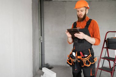 İnşaat işçisi, inşaat alanında dijital tablet kullanan kasklı sakallı bir adam. Tamirci ev tamiri yapıyor. Profesyonel müteahhit içeride. Boşluğu kopyala