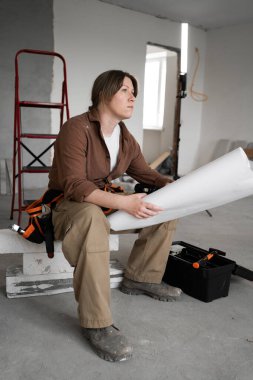DIY ve ev onarımı kavramı. Alet kemeri takan bir kadın odada oturuyor, yakınlardaki bir planlama evinde ya da apartmanda tadilat yapan bir alet kutusuyla kağıt planları okuyor.