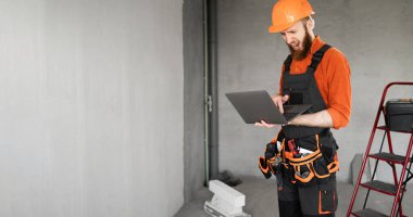 Meslek, inşaat ve inşaat kavramı. Sakallı erkek işçi ya da inşaatçı tulum giyiyor ve dizüstü bilgisayarı proje ya da planları yönetmek için kullanıyor. Pankart. Boşluğu kopyala