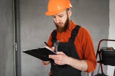 Meslek, inşaat ve inşaat kavramı. Sakallı bir erkek işçinin ya da inşaatçının portresi. Tulum giyip elinde pano tutuyor ve apartman yenileme projesi için notlar tutuyor..