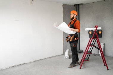 Sakallı miğferli ve tulumlu müteahhit dairenin planlarını merdivenle okuyor. Meslek, inşaat ve inşaat kavramı.