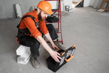 Meslek, inşaat ve inşaat kavramı. Sakallı erkek işçi ya da inşaatçı tulum giyip ev ya da apartman tadilatı yaparken profesyonel bir alet kutusunu açıyor.
