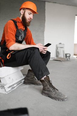 Meslek, inşaat ve inşaat kavramı. Sakallı erkek işçi ya da müteahhit tulum giyiyor ve kısa bir mola veriyor ve iş günü akıllı telefonunu iletişim için kullanıyor.