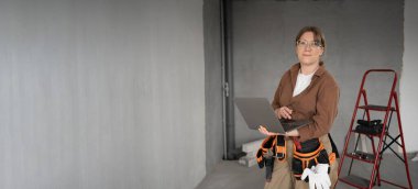 İşçi kadın yeni evdeki kameraya bakan dizüstü bilgisayarla alet kemeri takıyor. Yenileme dairesi. Evi ve teknoloji konseptini onar. Pankart. Boşluğu kopyala