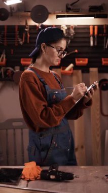 Mavi tulumlu kadın tamircinin küçük bir garajda ya da atölyede panoya yazı yazışının yan görüntüsü. dikey video