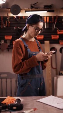 Küçük atölyedeki akıllı telefonları kullanarak internetten iş kıyafetleri ve şapka sohbeti yapan kadın tamirci. dikey video