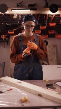 Marangozluk atölyesinde çalışırken gözlüklü ve kapak değiştiren profesyonel kadın marangoz. dikey video