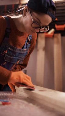 Profesyonel marangoz kadın marangozluk stüdyosunda aşındırıcı zımpara kağıdıyla kereste tahtasını cilalıyor. Gece ışığı altında atölyede manuel yüzey çalışması yapan katılımcı. dikey video