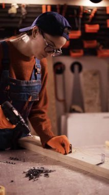 Gözlüklü ve tulumlu kadın marangoz otantik atölyedeki tahtadan vidayı sökmek için elektrikli matkap kullanıyor. dikey video