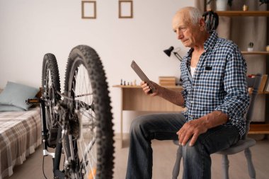 Evde dijital tablet kullanarak motosikletini kontrol eden ve tamir eden yaşlı bir adam. Üst düzey erkek bisikletçi bisiklete bakar. Boşluğu kopyala