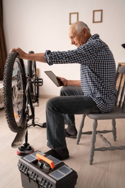 Evde dijital tablet kullanarak motosikletini kontrol eden ve tamir eden yaşlı bir adam. Üst düzey erkek bisikletçi bisiklete bakar. Boşluğu kopyala