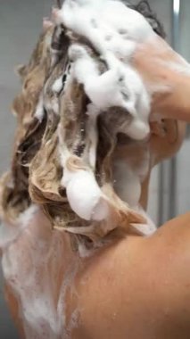 Duşta şampuanla uzun saçları yıkayan genç bir kadın. İlgi, güzellik ve sağlık konsepti. Dikey video