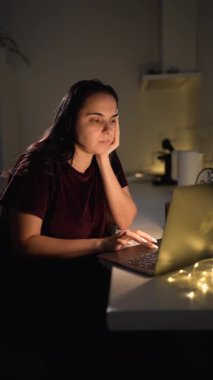 Pijamalı, sıkıcı genç bir kadın gece geç saatlere kadar mutfakta dizüstü bilgisayar başında çalışıyor. Fazla çalışma ve fazla mesai konsepti. dikey video