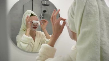 Banyoda aynanın yanında pamuk yüz maskesi takan genç bir kadın. Kadın yüz bakımı için kozmetik kullanıyor. Boşluğu kopyala