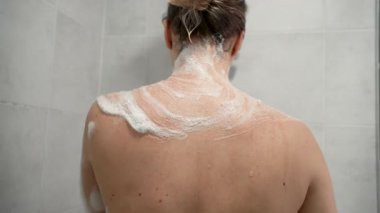 Duş alırken sırtını sünger ve sabun köpüğüyle ovuşturan genç bir kadının arka camı. Kişisel hijyen anlayışı, deri dökülmesi, vücut bakımı.