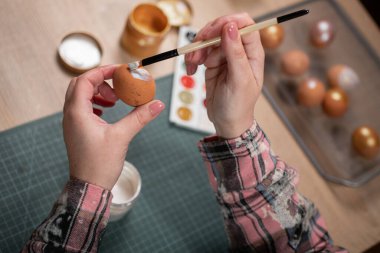 Paskalya yumurtaları evde. Paskalya yumurtasını fırça ve renkli palet kullanarak boyayan bir kadının yakın çekimi. Tatil yaratıcılığı ve şenlik hazırlıkları için rahat bir ortam..