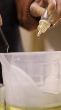 El yapımı mumlar. Kadın eli damlacığı mum yapımı için eritilmiş soya mumuna gerekli yağı katar. Yakın plan. dikey video