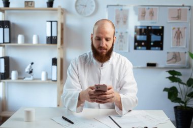 Beyaz önlüklü sakallı erkek doktor klinikte akıllı telefonuyla çalışıyor. Boşluğu kopyala