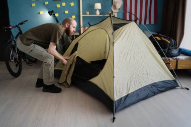 Sakallı adam, yürüyüş için hazırlanırken oturma odasının ortasına kamp çadırı kuruyor. Boşluğu kopyala