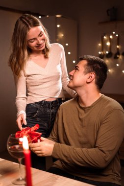 Sevgililer Günü 'nü evde romantik bir akşam yemeği yiyerek kutlayan mutlu genç çift. Sevgi dolu bir kadın erkeğine hediye kutusu veriyor. Boşluğu kopyala