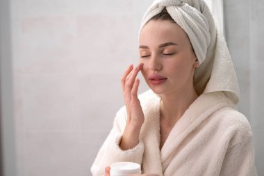 Genç, güzel bir kadın kafasında havluyla parlayan cildiyle yüzüne nemlendirici krem sürüyor. Cilt bakım, güzellik ve kozmetik konsepti
