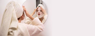 Bornozlu ve kafasında havlu olan genç bir kadın banyodaki aynaya bakarak nemlendirici çarşaf maskesi çıkarıyor. Pankart. Boşluğu kopyala
