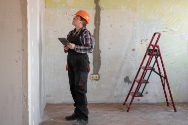 Tablet bilgisayar kullanan turuncu şapkalı bir kadın müteahhit veya işçi. İnşaat, yenileme ve teknoloji konsepti