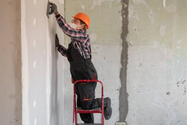 Sıva kaplı bir duvarı zımparalayan bir aletle kadın inşaatçı ya da tamirci. Ev tadilatı ve ev yenileme konsepti. Boşluğu kopyala
