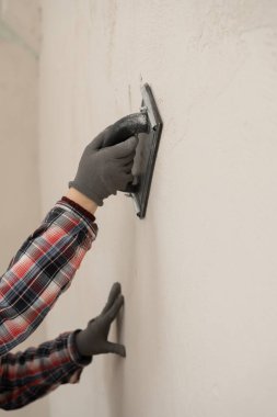 Duvar yüzeyini pürüzsüz hale getirmek için zımpara kağıdı kullanan inşaat işçisi, tadilat sırasında bitiriyor. yakın plan