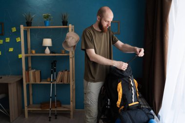 Sakallı yürüyüşçü eve dağ yolculuğu için sırt çantası hazırlıyor.