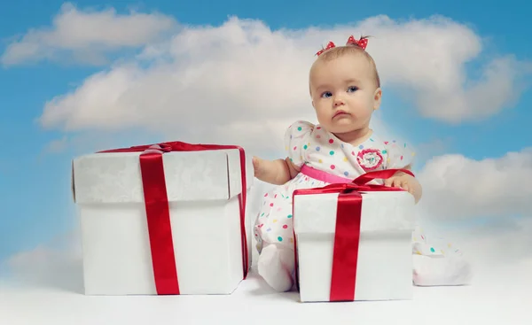 愛らしい赤ちゃんの女の子は 曇った空の背景の上にポーズをギフトボックスポイント指の脇に座っています 新年とクリスマスのお祝いのコンセプト — ストック写真