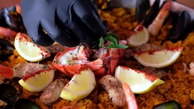 Ağır çekim video, paella geleneksel İspanyol mutfağını büyük, sığ bir tavada, siyah eldiven giymiş aşçının deniz ürünleri tabağını büyük ıstakoz ve otlarla süslemesi.
