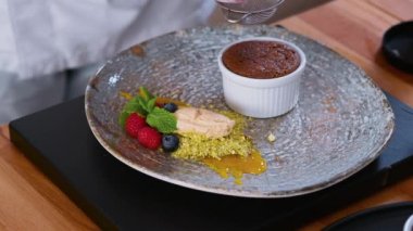 Şefin elleri üzerine pudra şekeri serpiştirip süzgeç süsleyerek dondurma, böğürtlen, sos ve şam fıstığı ile servis edilen çikolatalı kulan keki hazırlıyor. Restoran tatlı hazırlama konsepti