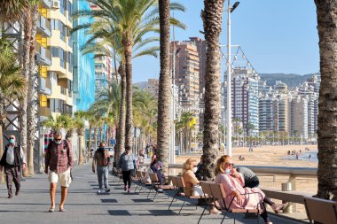 Benidorm, İspanya - 24 Aralık 2020: İnsanlar güneşli kış gününde Benidorm turistik kentinde deniz kenarında gezinti yapıyorlar. Costa Blanca 'da. Alicante vilayeti. İspanya
