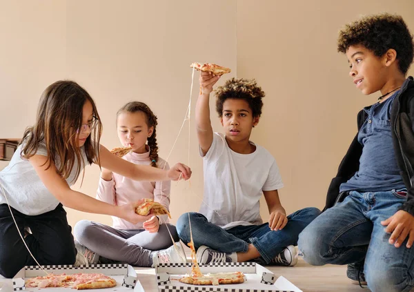 Group of pretty multi ethnic kids little boys girls sit indoor eating pizza italian fast food cuisine. Alpha generation share slices, spend time together. Friendship, time-to-eat lunch time concept