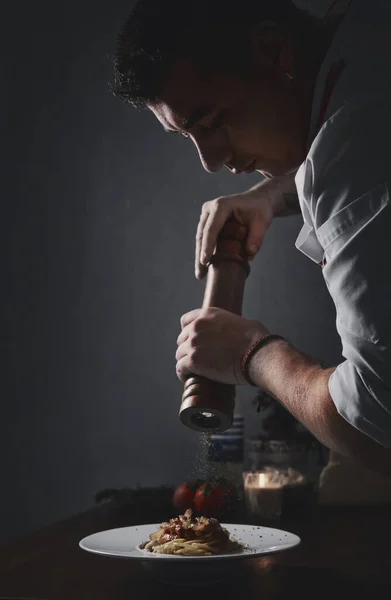 Side profile view young male chef holding pepper shaker peppering spaghetti. Restaurant main course dish, food preparation process concept