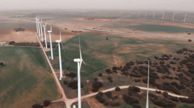 İHA açısı geniş. Yel değirmenlerinin hareket eden türbinlerinin havadan görüntüsü. Rüzgâr enerjisini elektriğe çevirir. Kasvetli bir günde ekilmiş çiftlik arazilerinin panoramik gerçek zamanlı videosu. Avrupa, İspanya