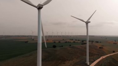İHA açısı geniş. Yel değirmenlerinin hareket eden türbinlerinin havadan görüntüsü. Rüzgâr enerjisini elektriğe çevirir. Kasvetli bir günde ekilmiş çiftlik arazilerinin panoramik gerçek zamanlı videosu. Avrupa, İspanya