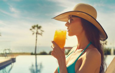 Side profile view face attractive woman, wear straw hat hold glass drinks natural orange juice non-alcoholic cocktail refreshing at hot day relax on deckchair near pool. Enjoy summer holidays concept