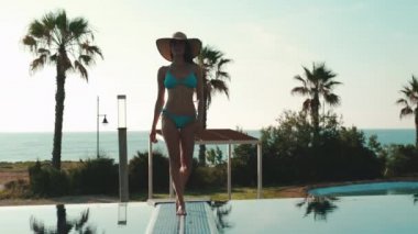 Bikinili uzun boylu, zayıf bir kadın, hasır şapkalı, güneş gözlüklü, yüzme havuzunun kenarından kameraya, palmiye ağaçlarına, arka planda deniz manzaralı. Yaz tatilleri, tropikal tatil konsepti.