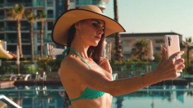Bikinili kadın, güneş gözlüğü, açık havuz kenarında poz veren hasır şapka akıllı telefon, yaz tatilinin özçekim fotoğrafını çek. Yaz tatili, modern teknoloji, güzellik konsepti